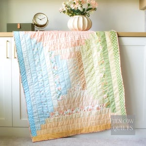 May include: A handmade quilt in a log cabin pattern, featuring pastel colors like peach, blue, and green. The quilt has a mix of solid and floral fabrics, with a dotted border. The text "TEN COW QUILTS" is visible in the lower right corner.