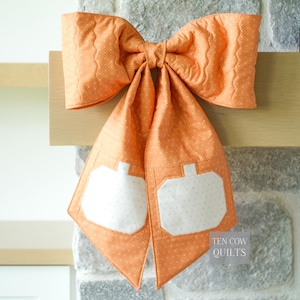 Puede incluir: Un lazo de tela acolchada en color naranja claro con un sutil patrón de puntos blancos. Las dos largas colas del lazo presentan aplicaciones blancas con forma de calabaza. El texto TEN COW QUILTS es visible en la esquina inferior derecha.