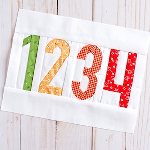 Peut inclure: Un bloc de tissu blanc avec les chiffres 1, 2, 3 et 4 appliqués en différents tissus à motifs. Le 1 est vert, le 2 est orange, le 3 est en vichy rouge et le 4 est rouge avec des motifs floraux blancs. Les chiffres sont cousus sur le fond blanc.