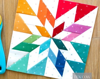 May include: A colorful quilt block with a starburst design. The block features a variety of colors including orange, red, pink, green, and blue. The background is white with a small cross pattern. The text "TEN COW QUILTS" is visible in the lower right corner.