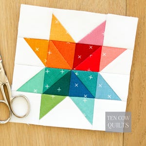Puede incluir: Un bloque de edredón colorido con un diseño de estrella. La estrella está compuesta por varios triángulos en tonos naranja, rojo, rosa, verde y azul, sobre un fondo blanco. Pequeñas cruces blancas están cosidas en los triángulos de colores. El texto "TEN COW QUILTS" está en la esquina inferior derecha.