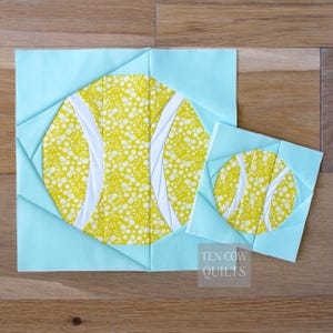 Peut inclure: Deux blocs de courtepointe en tissu avec un motif de balle de tennis. Les balles de tennis sont jaunes avec des rayures blanches et un motif floral. Les blocs sont sertis dans un tissu bleu clair. Le texte "TEN COW QUILTS" est visible.