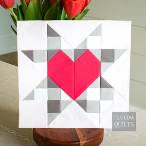 Könnte beinhalten: Ein quadratischer Quiltblock mit einem roten Herz in der Mitte, umgeben von einem Sternmuster in Grau und Weiß. Das Design hat eine moderne Ästhetik. Der Text "TEN COW QUILTS" ist unten rechts zu sehen. Ungefähr 30 cm x 30 cm.