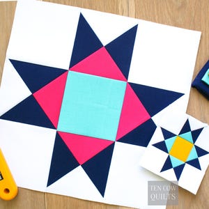 Puede incluir: Un gran bloque de edredón colorido con un diseño de estrella. La estrella presenta formas geométricas azul marino, rosa y turquesa sobre un fondo blanco. También hay un bloque más pequeño y similar. El texto "TEN COW QUILTS" es visible.