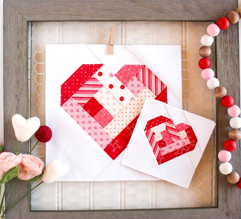 May include: Two white fabric squares with red and pink patchwork hearts. The hearts are made of various patterned fabrics with stripes, polka dots, and hearts. The squares are framed in a wooden frame.