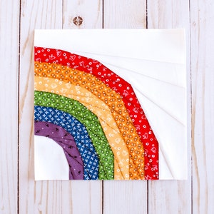 May include: A rainbow quilt block with a white background and colorful fabric strips in shades of red, orange, yellow, green, blue, and purple. The fabric strips are arranged in a curved shape, resembling a rainbow.