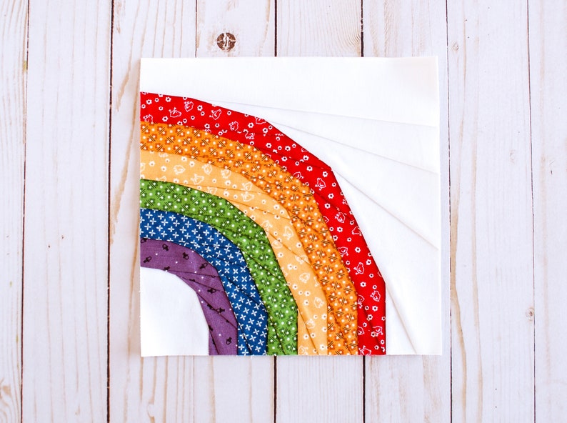 May include: A rainbow quilt block with a white background and colorful fabric strips in shades of red, orange, yellow, green, blue, and purple. The fabric strips are arranged in a curved shape, resembling a rainbow.