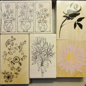 May include: A collection of wooden stamps featuring floral designs. The stamps include line drawings of flowers in vases, a rose, a bouquet, and a pink and yellow sunflower. The stamps are arranged in a rectangular shape.