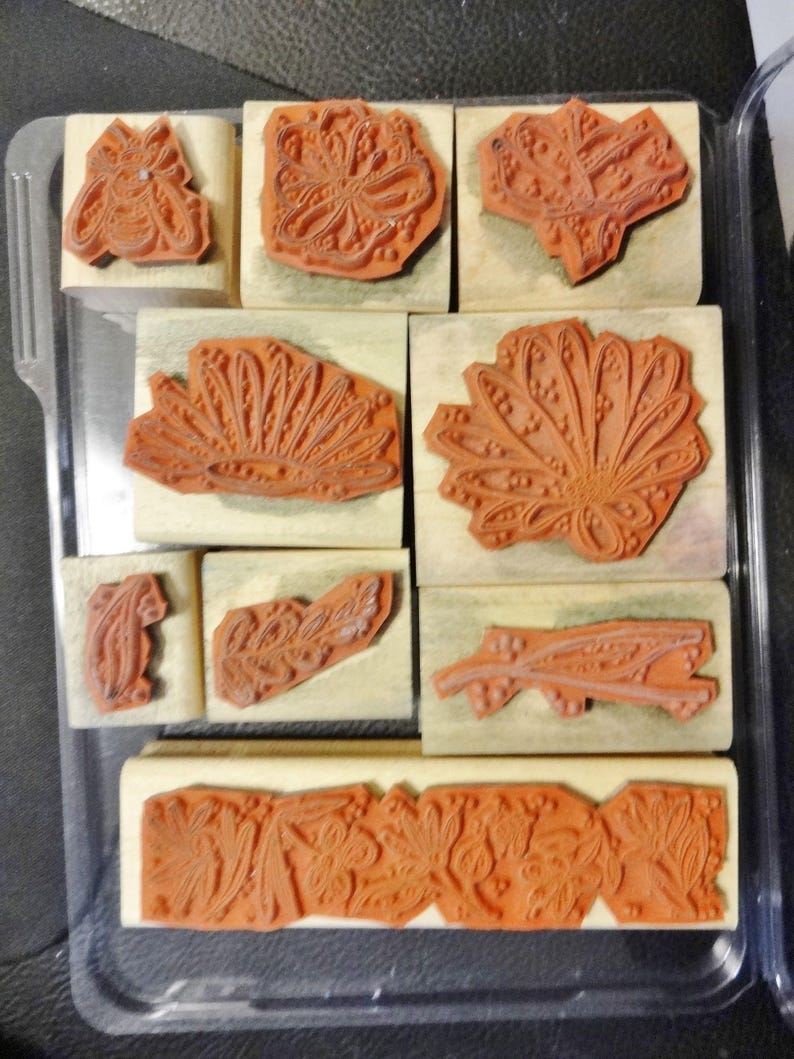 May include: A set of nine wooden mounted rubber stamps with floral designs. The stamps are arranged in a grid pattern in a clear plastic storage case.