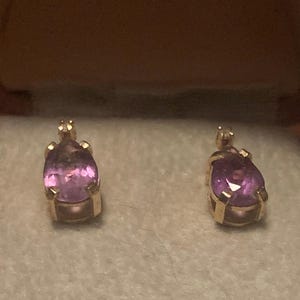 May include: Two gold-colored, pear-shaped earrings with a vibrant purple gemstone. The earrings are set in a classic bezel setting, showcasing the polished gemstones. The earrings are displayed on a light-colored, textured surface.