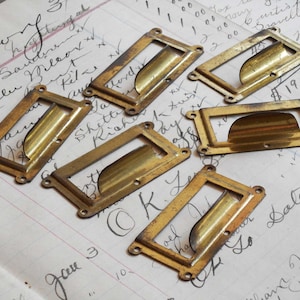 May include: Six rectangular brass label holders with a weathered, aged finish. Each holder features a central curved pull handle and four small circular screw holes at the corners for mounting, arranged in a scattered pattern on a paper surface.