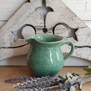 May include: A small, green ceramic pitcher with a handle and spout. The pitcher has a textured surface and sits on a wooden surface with dried lavender and greenery. A distressed, white wooden decorative element is in the background.