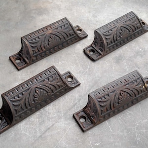 May include: Four dark brown metal drawer pulls featuring an ornate, raised floral and leaf pattern. Each handle has a curved, semi-cylindrical shape with flat mounting tabs on both ends, each containing a single circular screw hole for installation.