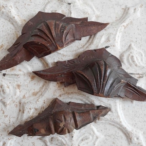 May include: Three antique, dark brown wooden leaf-shaped decorative elements. Each piece features detailed carving, with visible veins and textures. The leaves are arranged on a textured, off-white surface.