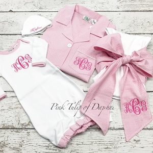 May include: A collection of baby clothing and accessories in white and pink seersucker fabric. Items include a gown, hat, pajamas, and a large bow, all with embroidered monograms. The hat is embroidered with the name "Kylee". The text "Pink Tulip of Daphne" is also visible.