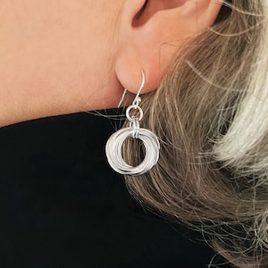 May include: A silver earring with a circular design, composed of multiple intertwined wires. The earring hangs from a simple hook, showcasing a modern and minimalist aesthetic. The jewelry is shown against a black background.