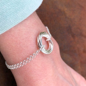 May include: A silver bracelet with a multi-ring design. The rings are intertwined and form a circle. The bracelet is on a person's wrist.