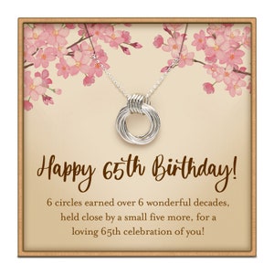 May include: A silver necklace with six interlocking circles, presented on a card with the text "Happy 65th Birthday!". The card features a floral design with pink blossoms and a brown border. The necklace is on a silver chain.