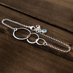 May include: A silver bracelet featuring three interconnected circles and a delicate chain. A small, round, blue gemstone is attached to the chain. The bracelet is displayed on a dark brown wooden surface.