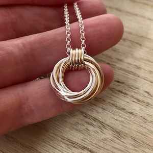 May include: A necklace featuring a pendant composed of multiple interlocking circular rings in alternating silver and gold tones. The pendant is suspended from a delicate silver-toned chain with round links, creating a layered, circular design.