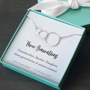 May include: A silver necklace with three interlocking circular rings in a gift box. The card reads: Three Generations, Grandmother, Mother, Daughter, three generations of awesome women! MAGGIE BLAKE JEWELRY, 925 STERLING SILVER. The box is teal with a white ribbon.