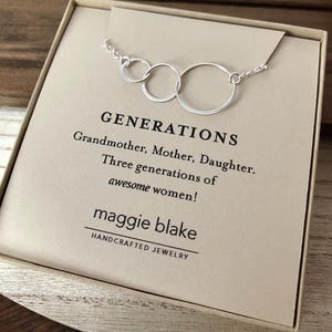 May include: A silver necklace with three interconnected circles, presented in a gift box. The card inside reads "GENERATIONS" and "Grandmother, Mother, Daughter. Three generations of awesome women!" The brand name "maggie blake" is also visible.