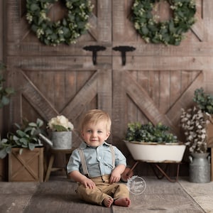 May include: A young boy wearing a light blue button-down shirt with suspenders and khaki pants sits on a wooden floor in front of a rustic wooden door backdrop. The door has two wreaths made of green leaves and there are various plants and wooden crates around the boy.
