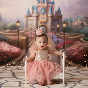 May include: A baby girl wearing a pink and gold sequined dress and a pink crown sits on a white miniature bed. The background is a blurred image of a fairytale castle with a path leading to it.
