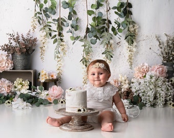 May include: A baby girl in a white dress with a lace collar sits in front of a white cake with white frosting and flowers. The background is a white wall with greenery hanging from the top.