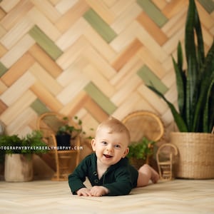 May include: A smiling baby wearing a green long-sleeved onesie lies on a wooden floor in front of a green and brown herringbone patterned wall.  The baby is surrounded by potted plants and wicker chairs.