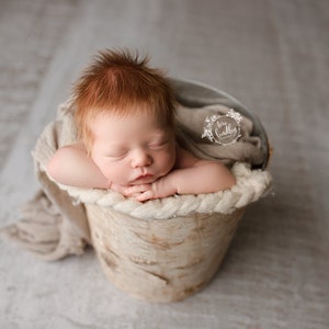 May include: A newborn baby with red hair is sleeping in a white birch bark bucket. The baby is wrapped in a beige blanket and is lying on its side with its head resting on its arm. The bucket is sitting on a light gray surface.