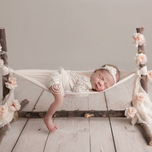 May include: A newborn baby sleeping in a white hammock made of fabric, decorated with pink flowers, and suspended between two wooden branches. The baby is wearing a white crocheted outfit and a white headband.