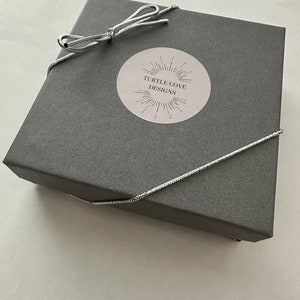 May include: A grey gift box with a silver ribbon tied in a bow. The box has a white sticker with the text "TURTLE COVE DESIGNS" and a sunburst design.