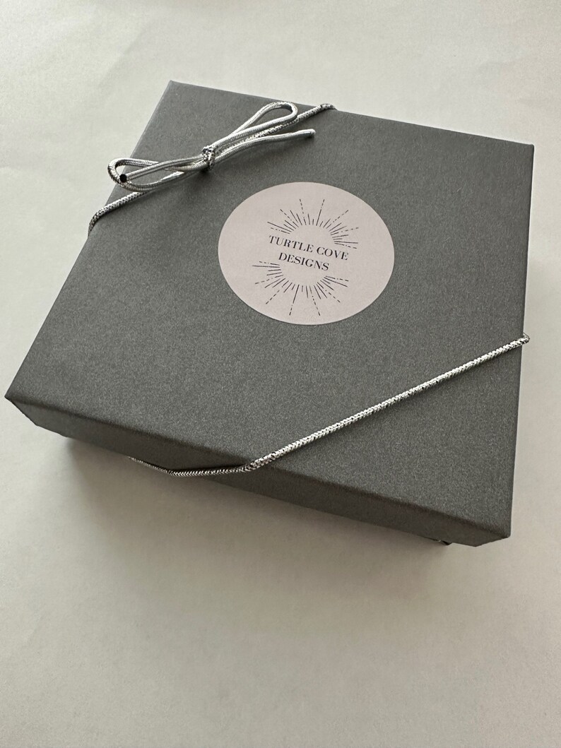 May include: A grey gift box with a silver ribbon tied in a bow. The box has a white sticker with the text "TURTLE COVE DESIGNS" and a sunburst design.