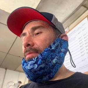 May include: A red and black baseball cap with a gray mesh back is worn by a person. A blue face covering with a floral and dragon pattern is also worn. The person is wearing a black shirt.
