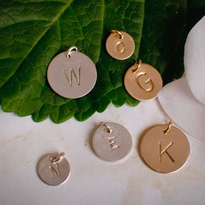 May include: Six round metal charms with different letters engraved on them. The charms are in gold and silver tones and are arranged on a white surface.