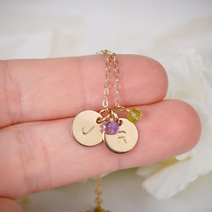 May include: Gold necklace with two small round charms, one with the letter 'J' and the other with the letter 'R'. The charms are attached to a delicate gold chain and have small purple and green gemstones.