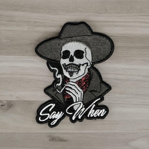 May include: Black and white embroidered patch of a skeleton wearing a cowboy hat and smoking a cigarette. The text "Say When" is below the skeleton.