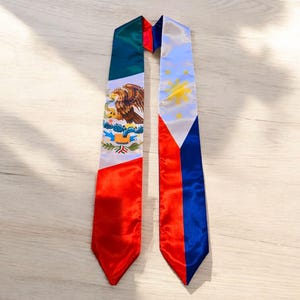 May include: A graduation sash with the flags of Mexico and the Philippines. The Mexican flag is green, white, and red with a golden eagle. The Philippine flag is blue, white, and red with a golden sun and three stars.