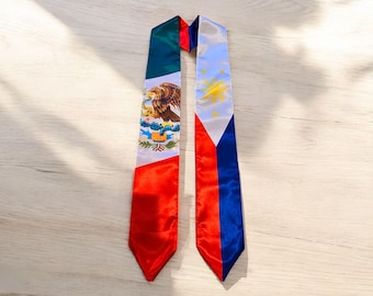 May include: A graduation sash with the flags of Mexico and the Philippines. The Mexican flag is green, white, and red with a golden eagle. The Philippine flag is blue, white, and red with a golden sun and three stars.