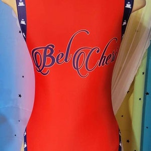 May include: Red bodysuit with navy blue trim and white stars. The text "Bel Cher" is printed on the back in a navy blue cursive font.