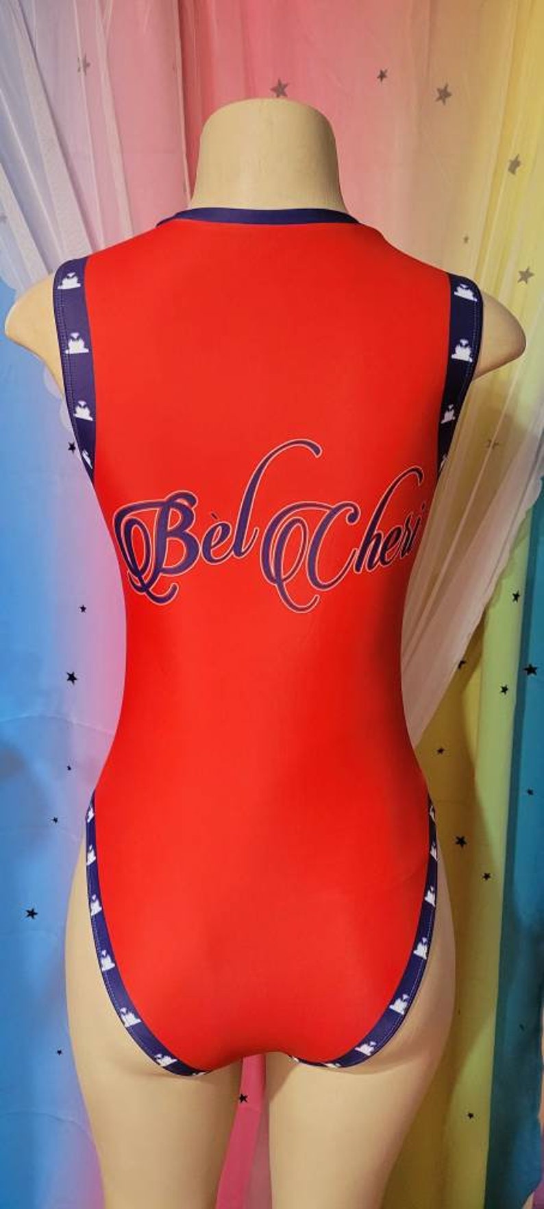 May include: Red bodysuit with navy blue trim and white stars. The text "Bel Cher" is printed on the back in a navy blue cursive font.