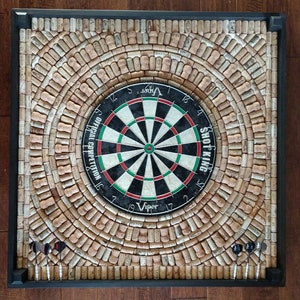 May include: Square dartboard with a black frame. The dartboard is surrounded by a mosaic of wine corks. The dartboard has a black, white, green, and red target design. Four darts are placed at the bottom.