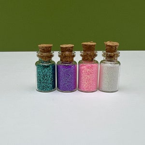 Puede incluir: Cuatro pequeños frascos de vidrio con tapones de corcho llenos de purpurina de diferentes colores. Los frascos están dispuestos en una fila de izquierda a derecha: azul verdoso, morado, rosa y blanco.