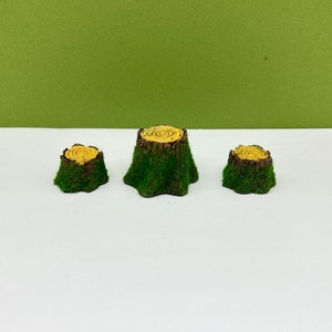 Könnte beinhalten: Drei Miniatur-Baumstümpfe, ideal für Feengärten oder Bastelprojekte. Die Stümpfe haben ein realistisches Aussehen mit einer moosgrünen Basis und einer hellbraunen Oberseite mit konzentrischen Kreisen. Der Hintergrund ist einfarbig grün.