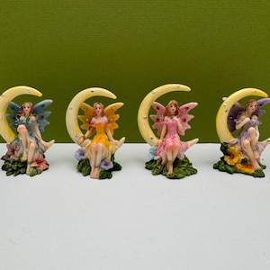 May include: Four fairy figurines, each seated on a yellow crescent moon. The fairies wear dresses in blue, yellow, and pink, with matching wings. Each figurine is surrounded by colorful flowers and greenery, creating a whimsical scene.