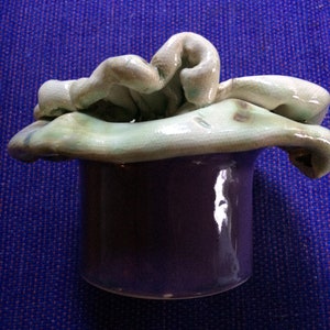 May include: A ceramic vase with a deep purple body and a light green, textured, organic-shaped top. The vase has a glossy finish and sits on a blue textured surface. The top appears to be a stylized flower or abstract design.