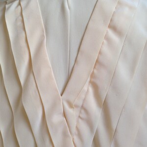 Op de afbeelding: Close-up van een cr&egrave;mekleurige blouse. De achterkant van de blouse heeft een V-hals met gelaagde stoffen details. Een kleine, ronde, witte knoop sluit de bovenkant van de blouse. De stof heeft een subtiele glans.