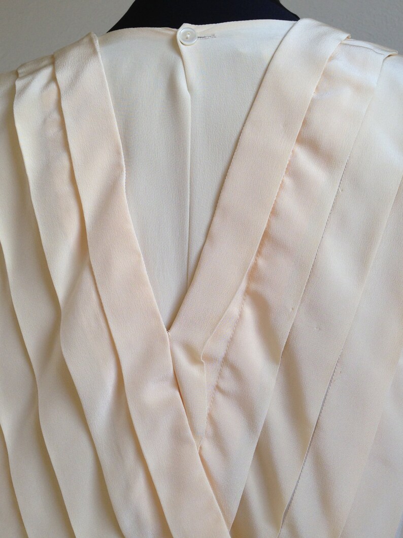 Op de afbeelding: Close-up van een cr&egrave;mekleurige blouse. De achterkant van de blouse heeft een V-hals met gelaagde stoffen details. Een kleine, ronde, witte knoop sluit de bovenkant van de blouse. De stof heeft een subtiele glans.