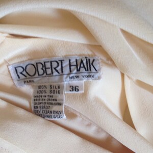 Op de afbeelding: Close-up van een cr&egrave;mekleurig zijden kledingstuk met een label met de tekst "ROBERT HAIK PARIS NEW YORK". Het label geeft aan dat het kledingstuk 100% zijde is, maat 36, en gemaakt in Hong Kong.